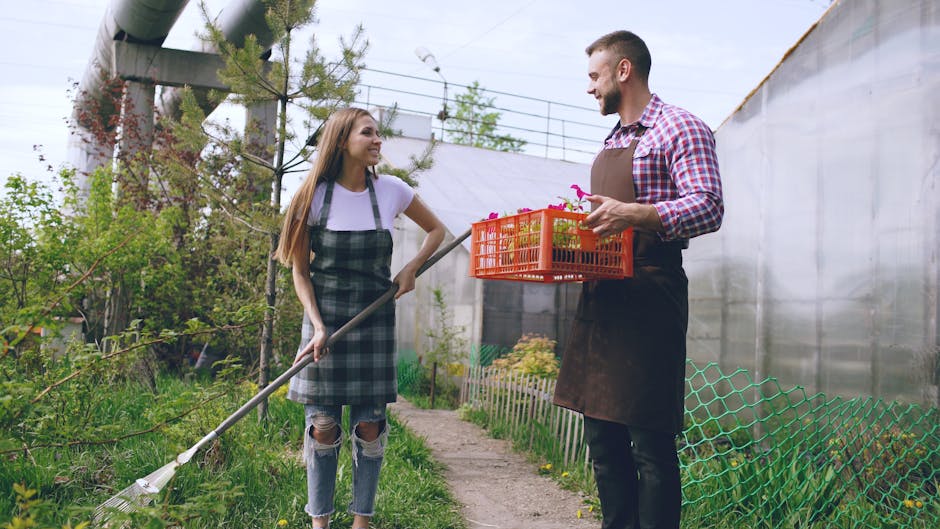 A young woman in a casual outfit, including a white t-shirt and ripped jeans, is holding a garden rake and smiling while engaging in conversation with a man dressed in a checkered shirt and a dark apr