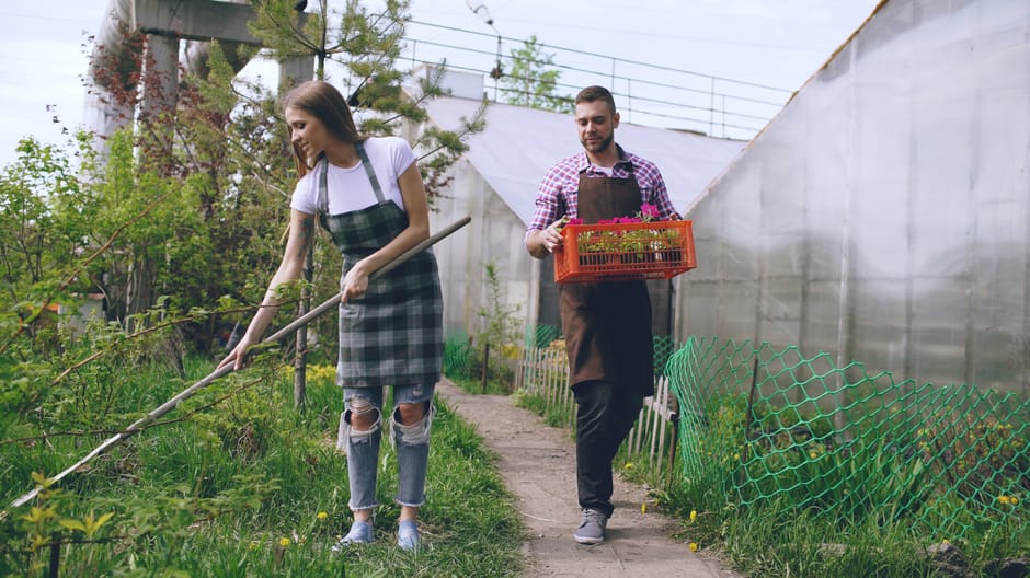A young woman in a casual outfit, including a white t-shirt and ripped jeans, is holding a garden rake and smiling while engaging in conversation with a man dressed in a checkered shirt and a dark apr
