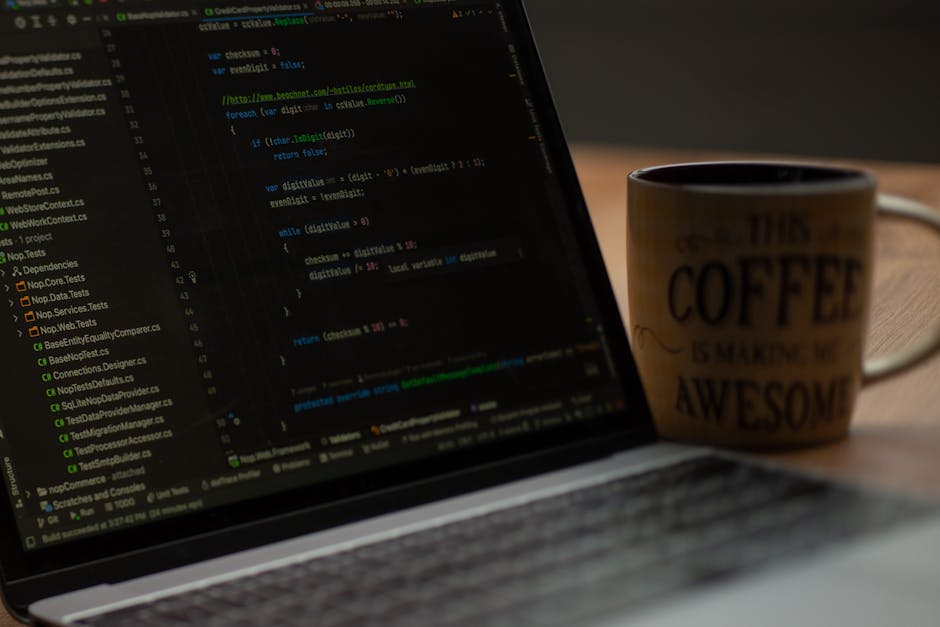 A close-up image of a laptop computer displaying lines of green, blue, and purple code on a black background, placed on a wooden desk. Next to the laptop, there is a ceramic mug with an off-white back
